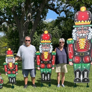 May include: Four nutcracker figurines of varying sizes, standing on a grassy lawn. The largest nutcracker is red and green with a gold crown and a white beard. The other nutcrackers are smaller and have similar designs.