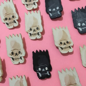 May include: A collection of wooden skull pins in various shades of brown and black. The skulls are painted with a cartoonish style and have a spiky hair design.
