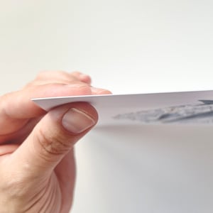 May include: A hand holding a white paper print with a grayscale illustration. The print's edge is visible, showing its thickness. The background is plain white.