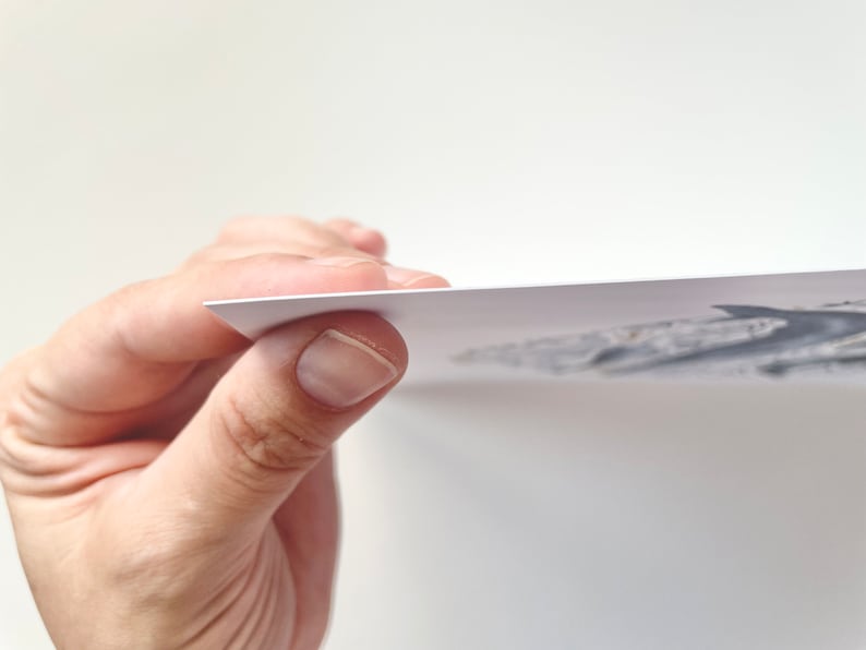 May include: A hand holding a white paper print with a grayscale illustration. The print's edge is visible, showing its thickness. The background is plain white.