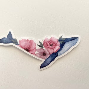 Könnte beinhalten: Ein Aquarell-Aufkleber einer Wal mit dunkelblauem Körper und Schwanz, verziert mit rosa und lila Blumen und grünen Blättern. Der Aufkleber befindet sich auf einem weißen Hintergrund.