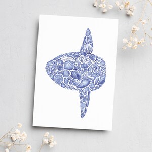 Peut inclure: Une carte blanche avec une illustration bleue d'un poisson-lune composée de coquillages. Le motif du poisson-lune est détaillé avec différentes formes et textures de coquillages, créant un thème marin. La carte est posée sur un fond gris clair avec de délicates fleurs blanches.