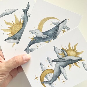 May include: Three white art prints featuring watercolor illustrations of whales, suns, moons, clouds, and stars. The whales are gray, the suns and moons are gold, and the clouds are gray and white.