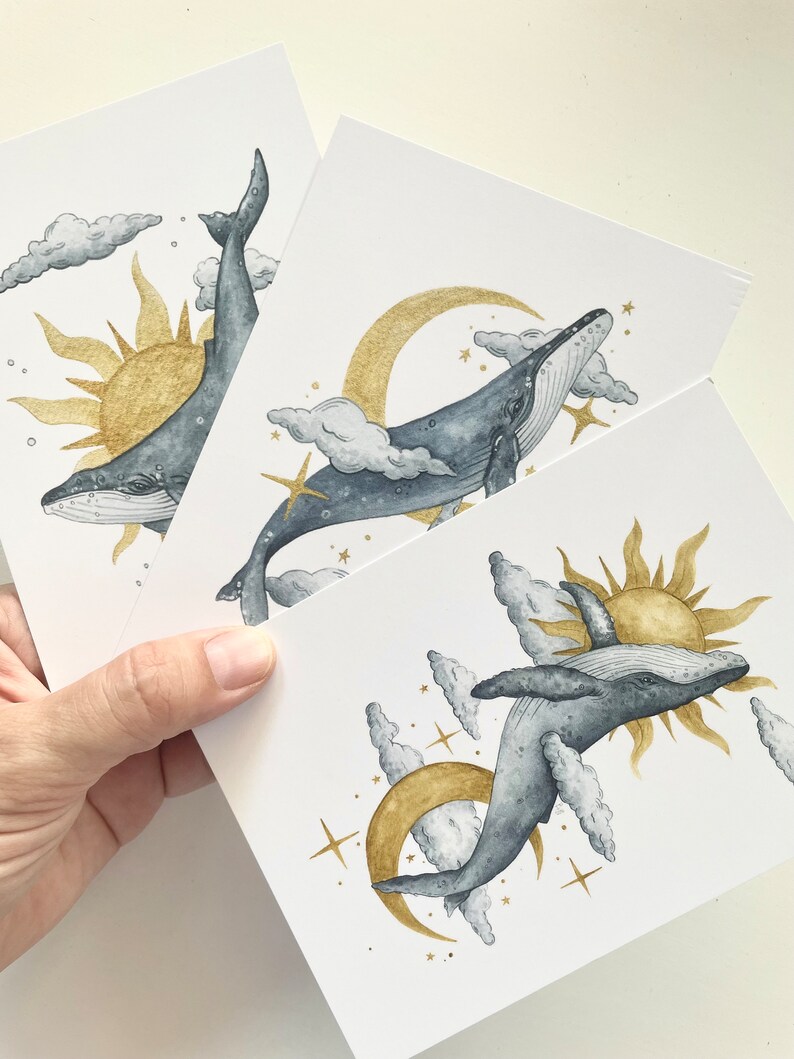 May include: Three white art prints featuring watercolor illustrations of whales, suns, moons, clouds, and stars. The whales are gray, the suns and moons are gold, and the clouds are gray and white.