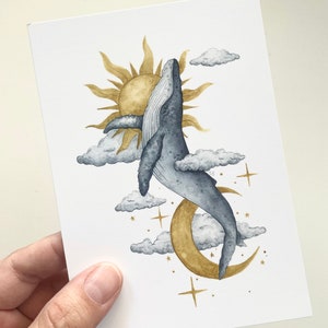 May include: Watercolor art print featuring a whale leaping towards a golden sun and moon. The artwork includes fluffy clouds and gold stars against a white background. The whale is depicted in shades of blue and gray.