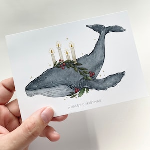 Può includere: Biglietto di Natale ad acquerello con una balena grigia adornata da una ghirlanda di agrifoglio e bacche, con quattro candele accese. Il testo "WHALEY CHRISTMAS" è stampato in basso.