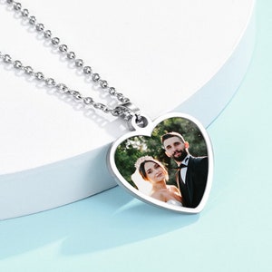May include: A silver heart-shaped pendant necklace with a photo of a couple in the centre. The necklace is on a white surface.