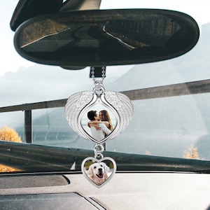 May include: A silver car charm with a heart-shaped frame featuring a photo of a couple. The heart is attached to a pair of angel wings. A smaller heart-shaped frame with a photo of a dog is hanging below the larger heart.
