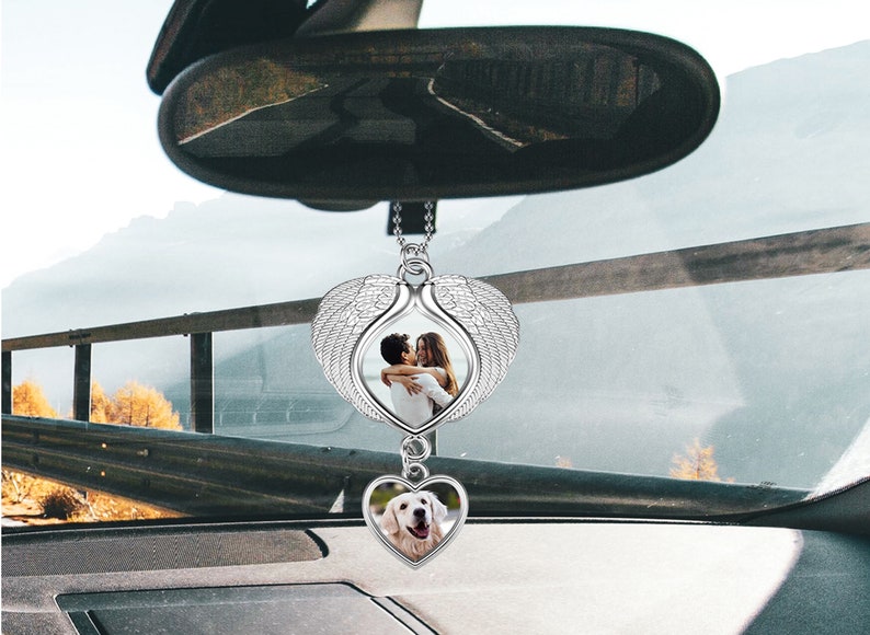 May include: A silver car charm with a heart-shaped frame featuring a photo of a couple. The heart is attached to a pair of angel wings. A smaller heart-shaped frame with a photo of a dog is hanging below the larger heart.