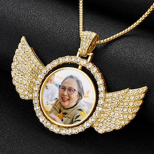 Op de afbeelding: Gouden ketting met hanger met een fotomedaillon met een lachende vrouw. Het medaillon is omringd door heldere strass steentjes en heeft aan beide kanten twee vleugels, die ook met strass steentjes zijn versierd.
