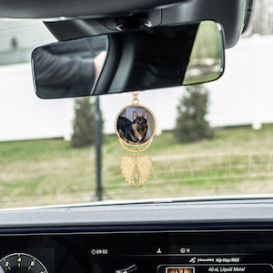 May include: Gold car charm with a round photo of a dog and gold angel wings. The charm is hanging from the rearview mirror of a car.