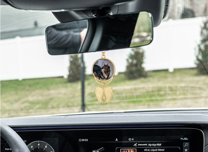May include: Gold car charm with a round photo of a dog and gold angel wings. The charm is hanging from the rearview mirror of a car.