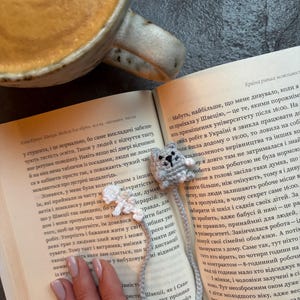 May include: A handmade gray crocheted bookmark featuring a small, smiling cat head and a white flower. The bookmark is placed on an open book with text, next to a cup of coffee.