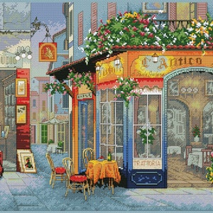 Puede incluir: Un patrón de punto de cruz que representa una encantadora escena de calle italiana con una cafetería llamada "L'Antico Sigillo" con un toldo floral, una fachada azul y blanca y una mesa con sillas para dos.