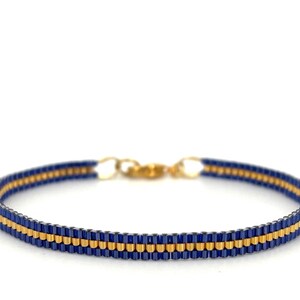 Op de afbeelding: Een kralenarmband met een gouden sluiting. De armband heeft een patroon van afwisselende rijen marineblauwe en gouden kralen. De armband is ongeveer 1,3 cm breed en staat tegen een witte achtergrond.