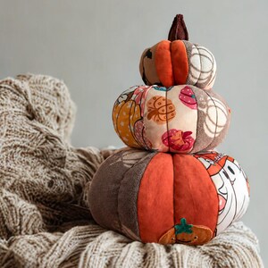 May include: A stack of three plush fabric pumpkins in varying sizes. The pumpkins feature a patchwork design with orange, brown, and cream colours, decorated with patterns of small pumpkins, stars, and ghost figures. A small brown stem sits on top.