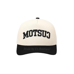 Könnte beinhalten: Cremefarbene Baseballkappe mit schwarzem Schirm und dem Wort "MOTSUJ" in Schwarz gestickt. Der Text "Made by Us Boutique" befindet sich unter dem Schirm. Die Kappe hat einen gebogenen Schirm und einen Knopf oben.