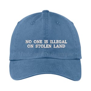 May include: A denim blue baseball cap with a curved peak. The cap has white embroidered text: "NO ONE IS ILLEGAL ON STOLEN LAND". The hat is a classic six-panel design, suitable for casual wear.