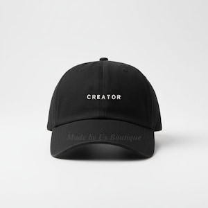 Puede incluir: Gorra de béisbol negra con la palabra "CREATOR" en letras blancas. La gorra tiene una visera curva y una corona redondeada. El texto "Made by Us Boutique" está impreso en la parte inferior de la gorra.