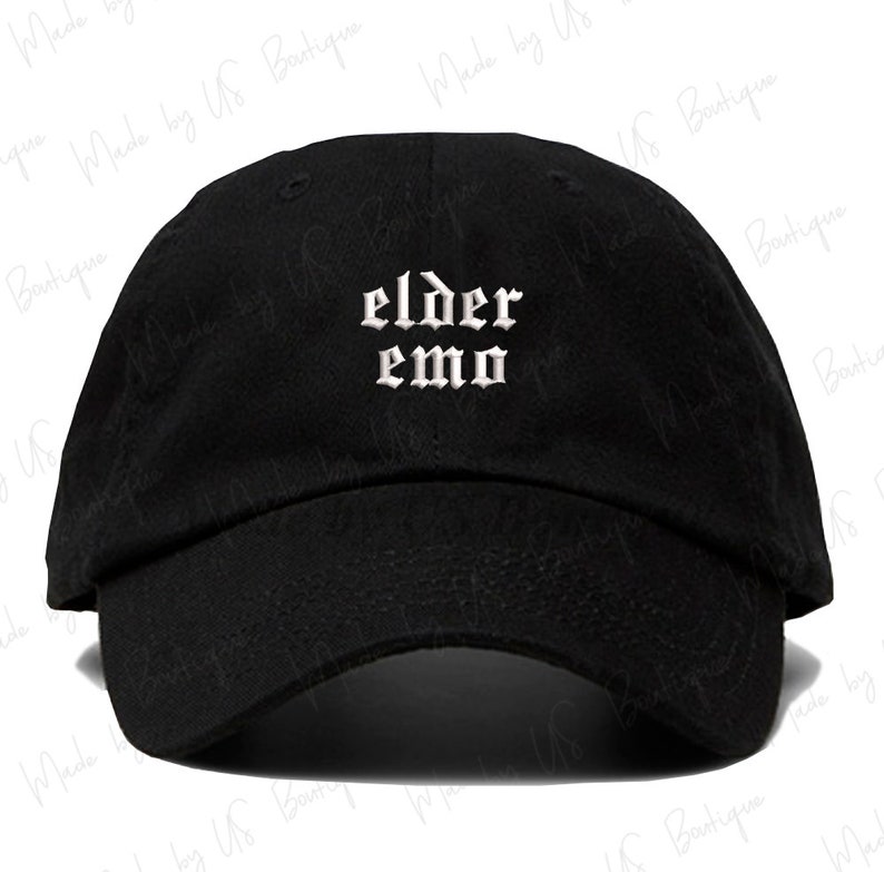 The image shows a black cap with the words "elder emo" embroidered in a gothic font. It is held outdoors, with a background of green grass, trees, and a small body of water under a partly cloudy sky, creating a peaceful atmosphere.
