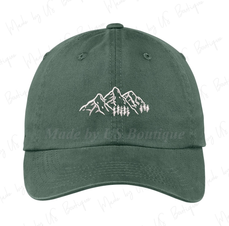 The image shows a green cap with white embroidered mountains and trees. It's held over a grassy background outdoors, complementing the nature-inspired design. The setting feels serene, with the cap evoking an outdoorsy vibe.