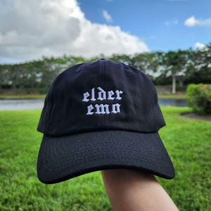The image shows a black cap with the words "elder emo" embroidered in a gothic font. It is held outdoors, with a background of green grass, trees, and a small body of water under a partly cloudy sky, creating a peaceful atmosphere.