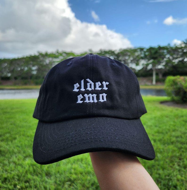 The image shows a black cap with the words "elder emo" embroidered in a gothic font. It is held outdoors, with a background of green grass, trees, and a small body of water under a partly cloudy sky, creating a peaceful atmosphere.