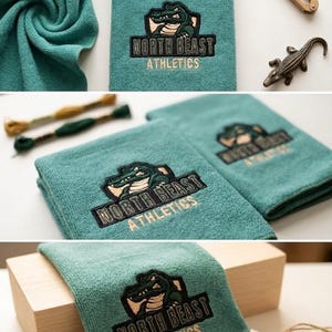 以下が含まれることがあります： 「NORTH BEAST ATHLETICS」の文字とワニのロゴが刺繍された、ターコイズ色のタオル。タオルは折りたたまれ、小さなアリゲーターの置物とハサミと一緒に展示されています。