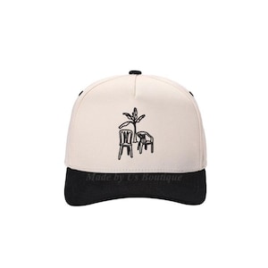 Puede incluir: Gorra de béisbol color crema con visera negra. La gorra presenta un diseño bordado en negro de una silla, una mesa y una planta. El texto "Made by Us Boutique" está impreso en la visera.