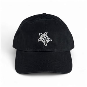 Pode incluir: Boné de beisebol preto com um design de tartaruga marinha bordada em branco. O texto "MadebyUsBoutique" é bordado na parte frontal do boné.
