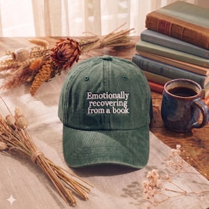 Może przedstawiać: Zielona czapka bejsbolowa z białym haftem "Emotionally recovering from a book". Na drewnianym stole, z książkami i kubkiem kawy. Suszone kwiaty i neutralna tkanina dopełniają scenę.