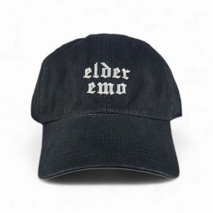 The image shows a black cap with the words "elder emo" embroidered in a gothic font. It is held outdoors, with a background of green grass, trees, and a small body of water under a partly cloudy sky, creating a peaceful atmosphere.