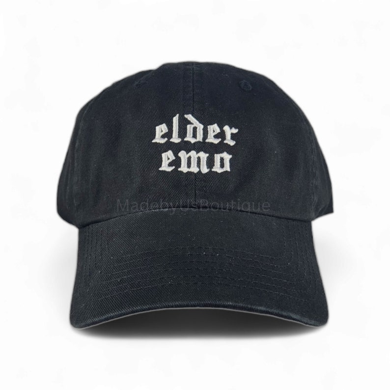 The image shows a black cap with the words "elder emo" embroidered in a gothic font. It is held outdoors, with a background of green grass, trees, and a small body of water under a partly cloudy sky, creating a peaceful atmosphere.