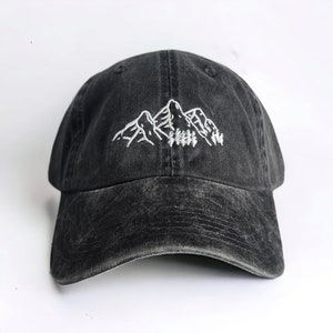 The image shows a washed black cap with white embroidered mountains and trees. It's held over a grassy background outdoors, complementing the nature-inspired design. The setting feels serene, with the cap evoking an outdoorsy vibe.
