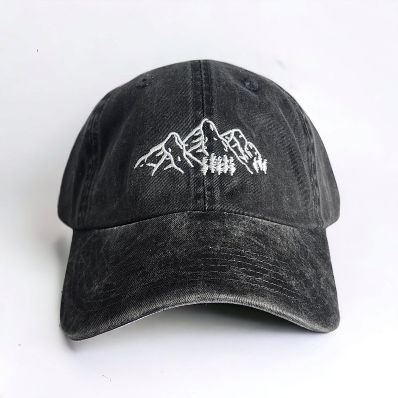 The image shows a washed black cap with white embroidered mountains and trees. It's held over a grassy background outdoors, complementing the nature-inspired design. The setting feels serene, with the cap evoking an outdoorsy vibe.