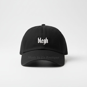 May include: A black baseball cap with the word "blegh" in white gothic font across the front. The cap has a curved brim and a rounded crown. The text "Made by Us Boutique" is printed in white on the brim.