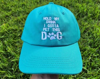May include: A turquoise blue baseball cap with white embroidery that reads "Hold my drink I gotta pet this D.O.G." with a paw print in place of the "O".