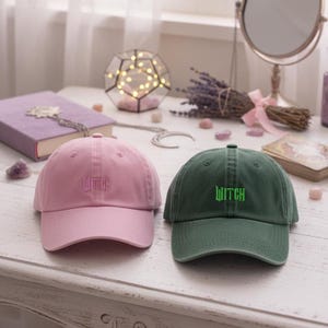 May include: Two baseball caps, one in dusty rose and one in dark green. Each cap has the word "WITCH" embroidered on the front. The dusty rose cap has matching embroidery, while the dark green cap has green embroidery. The caps are on a white surface.