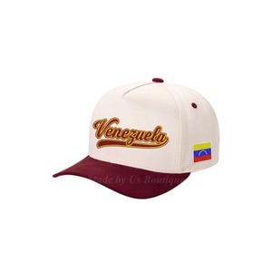 May include: Cream-colored baseball cap with a maroon brim and top button. The word "Venezuela" is embroidered in a gold script across the front. A small Venezuelan flag patch is on the side.