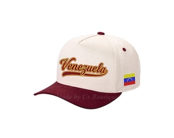 Può includere: Cappellino da baseball color crema con visiera e bottone bordeaux. La parola "Venezuela" è ricamata in oro sul davanti. Una piccola toppa con la bandiera venezuelana è sul lato.