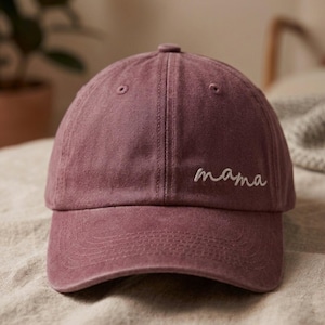 May include: A dusty rose baseball cap with the word "mama" embroidered in white script across the front. The cap has a curved brim and a classic six-panel design. The hat is made of a soft, textured fabric.