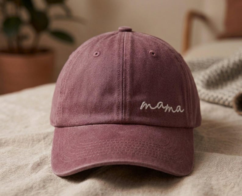 May include: A dusty rose baseball cap with the word "mama" embroidered in white script across the front. The cap has a curved brim and a classic six-panel design. The hat is made of a soft, textured fabric.