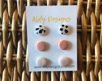 May include: A set of six stud earrings on a white card with the text "Aldy Designs" and the website "www.aldydesigns.etsy.com". The earrings are in three different designs: cow print, solid peach, and white with glitter.