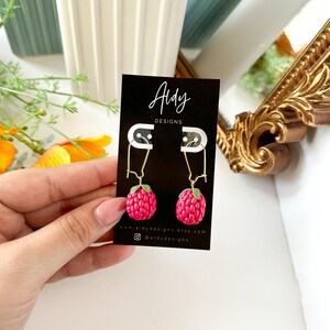 Op de afbeelding: Een paar oorhangers met roze framboosvormige bedels met groene bladeren. De oorhangers bevinden zich op een witte kaart met de tekst "Aidy Designs" en website-informatie.
