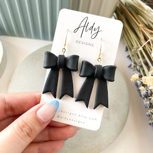 Puede incluir: Pendientes de lazo negro con ganchos dorados. Los pendientes están en una tarjeta blanca con el texto "Aldy Designs" y la dirección del sitio web "aldydesigns.etsy.com".