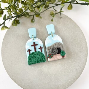 K&ouml;nnte beinhalten: Ein Paar handgefertigte Ohrringe aus Polymer Clay mit religi&ouml;sem Thema. Ein Ohrring zeigt drei Kreuze auf einem gr&uuml;nen H&uuml;gel, w&auml;hrend der andere ein Steingrab mit einem rollenden Stein zeigt.