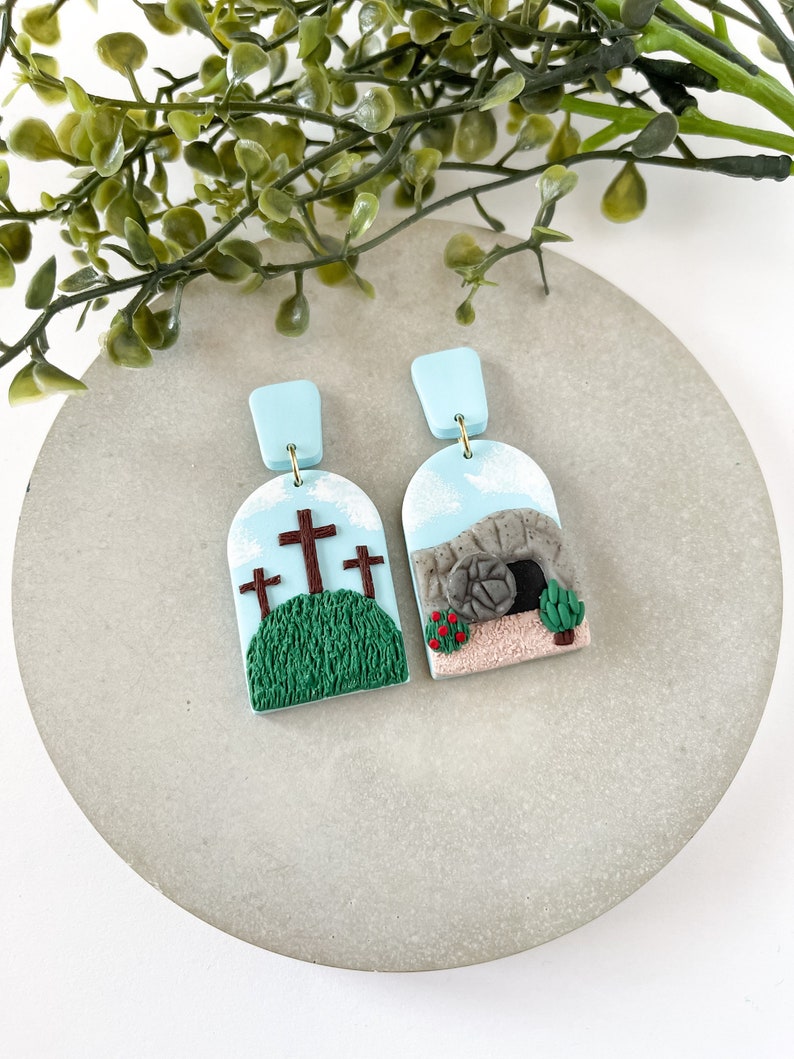 K&ouml;nnte beinhalten: Ein Paar handgefertigte Ohrringe aus Polymer Clay mit religi&ouml;sem Thema. Ein Ohrring zeigt drei Kreuze auf einem gr&uuml;nen H&uuml;gel, w&auml;hrend der andere ein Steingrab mit einem rollenden Stein zeigt.