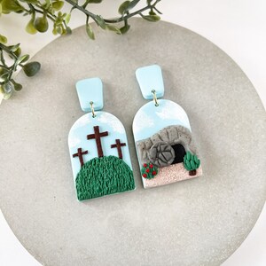 K&ouml;nnte beinhalten: Ein Paar blaue und wei&szlig;e Ohrringe aus Ton mit religi&ouml;sem Motiv. Ein Ohrring zeigt drei braune Kreuze auf einem gr&uuml;nen H&uuml;gel mit blauem Himmel. Der andere Ohrring zeigt ein graues Steingrab mit einer braunen T&uuml;r und einer roten Blume.