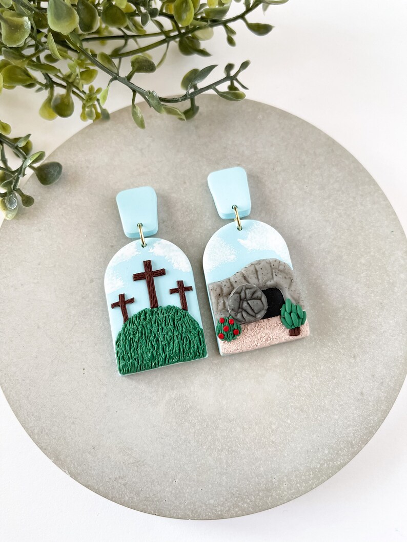 K&ouml;nnte beinhalten: Ein Paar blaue und wei&szlig;e Ohrringe aus Ton mit religi&ouml;sem Motiv. Ein Ohrring zeigt drei braune Kreuze auf einem gr&uuml;nen H&uuml;gel mit blauem Himmel. Der andere Ohrring zeigt ein graues Steingrab mit einer braunen T&uuml;r und einer roten Blume.