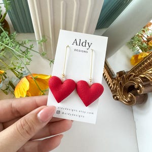 Op de afbeelding: Een paar rode hartvormige oorbellen met gouden haakjes. De oorbellen worden getoond op een witte kaart met de tekst "Aldy Designs" en de social media handle "@aldydesigns".